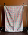 Semi-antique Indian quilt with a pink and mint green geometric triangle and diamond pattern hanging on a clothing rack against a terracotta wall.