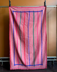 Semi-antique Indian quilt with a warm pink base, contrasting blue border frame, and vertical red and green stripes hanging on a rack against a dark wall.