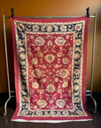 Semi-antique Indian quilt with a deep rust red base, ornate cream floral motifs, and a dark blue patterned border hanging on a rack against a brown wall.