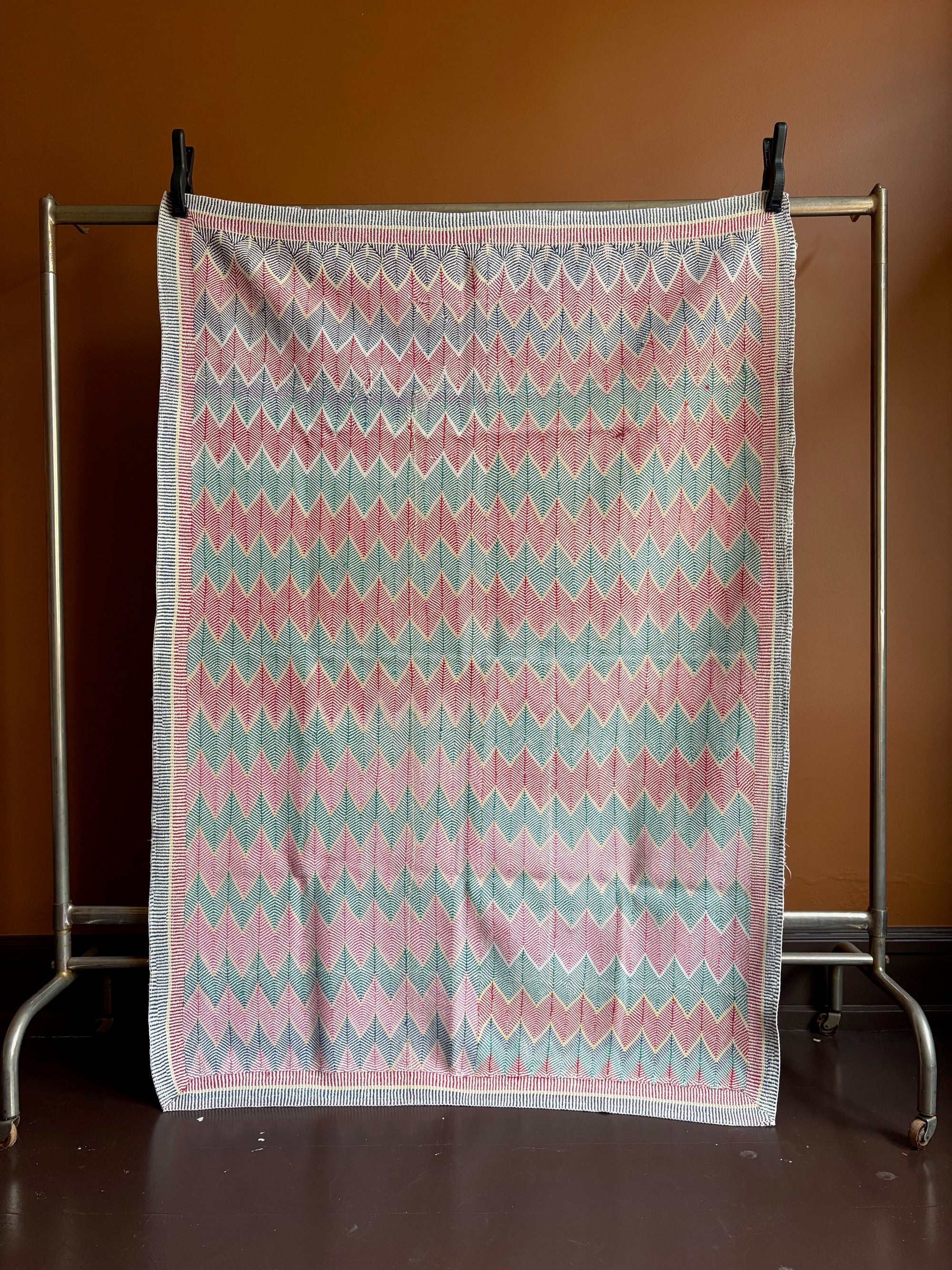 Semi-antique Jaipur Kantha quilt with a pink and mint green chevron zig-zag pattern hanging on a clothing rack against a terracotta wall.