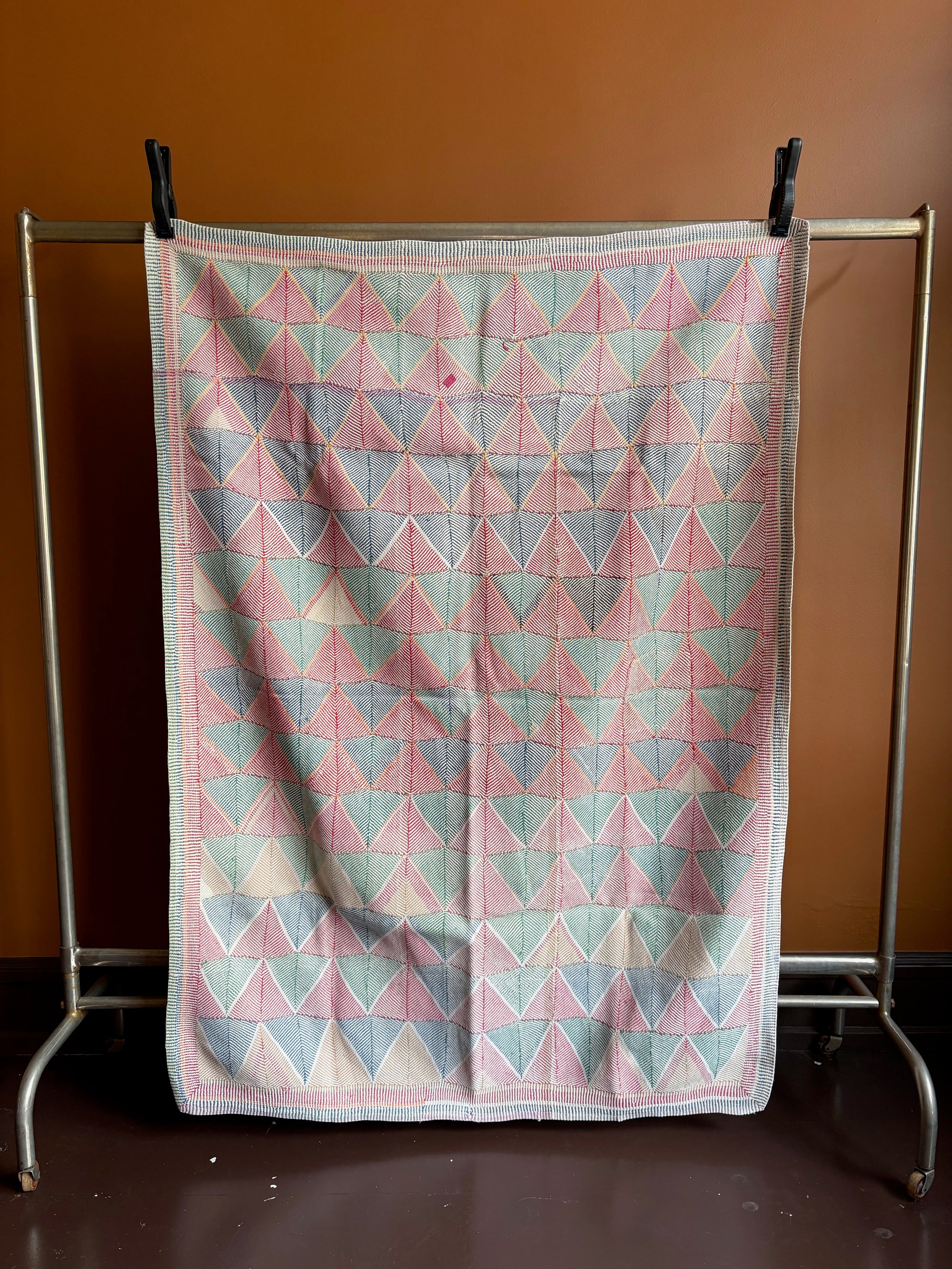 Semi-antique Indian quilt with a pink and mint green geometric triangle and diamond pattern hanging on a clothing rack against a terracotta wall.