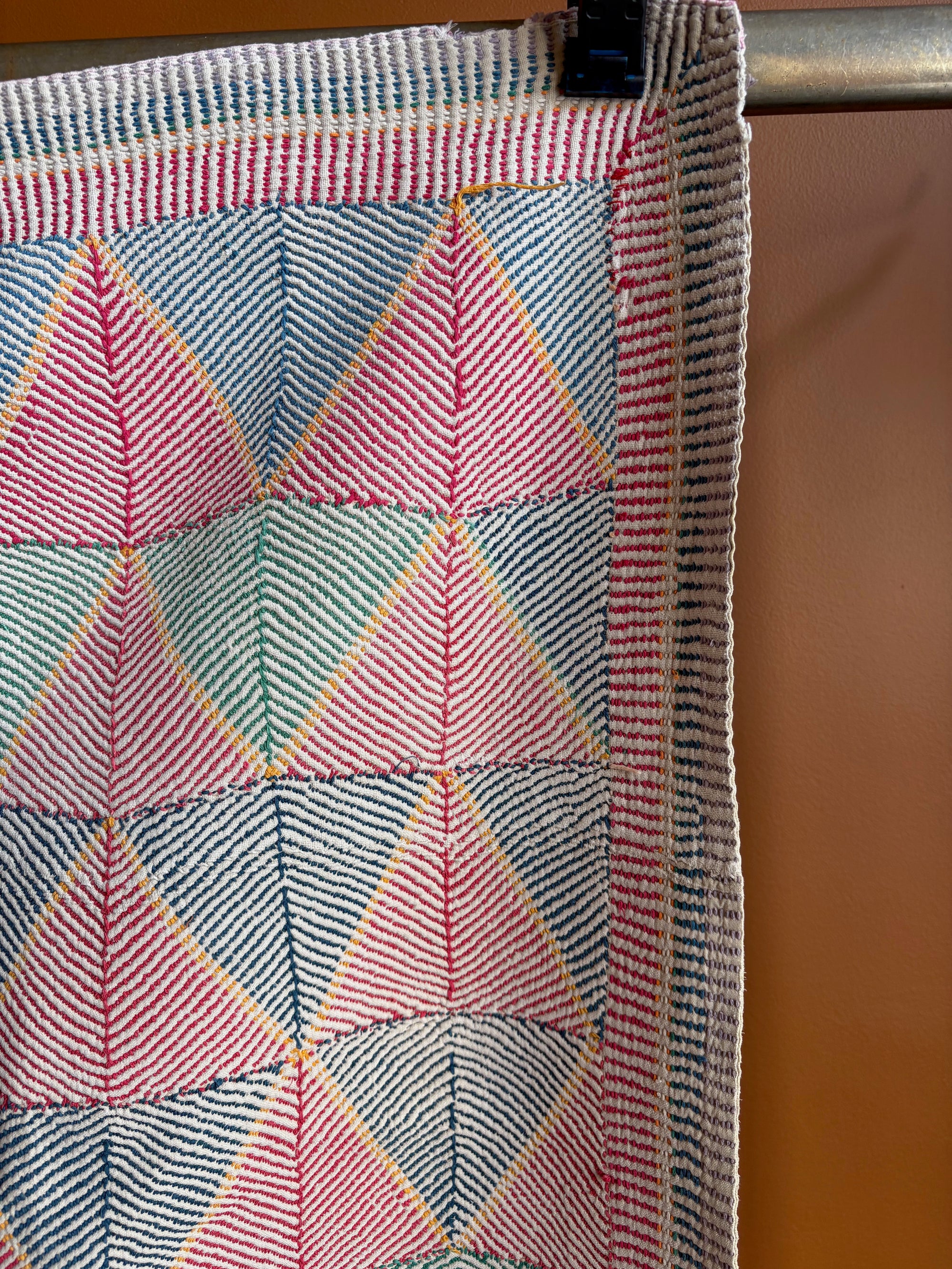 Semi-antique Indian quilt with a pink and mint green geometric triangle and diamond pattern hanging on a clothing rack against a terracotta wall.