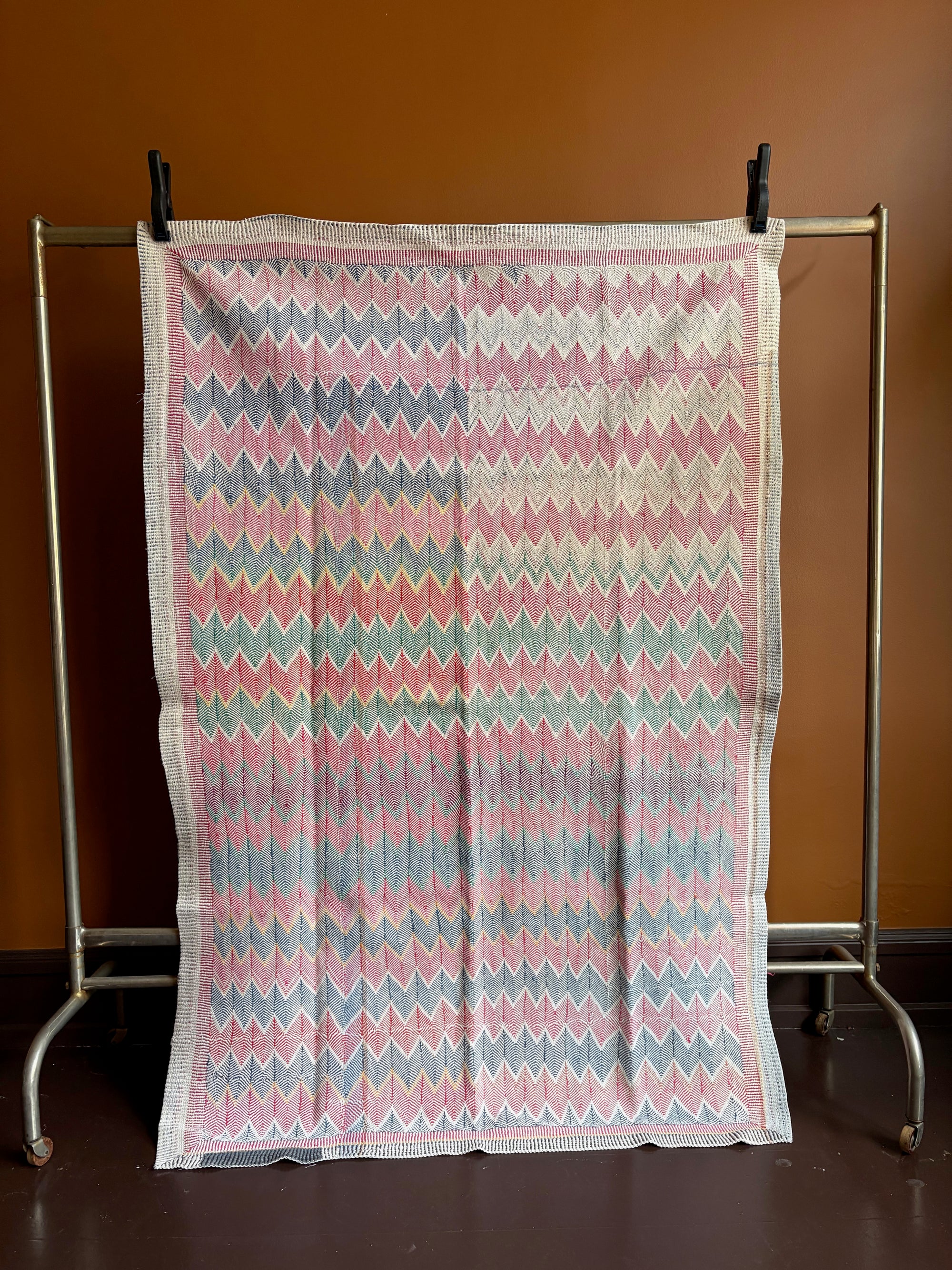 Semi-antique Jaipur Kantha quilt with a two-tone indigo blue and red chevron zig-zag pattern hanging on a clothing rack against a terracotta wall.