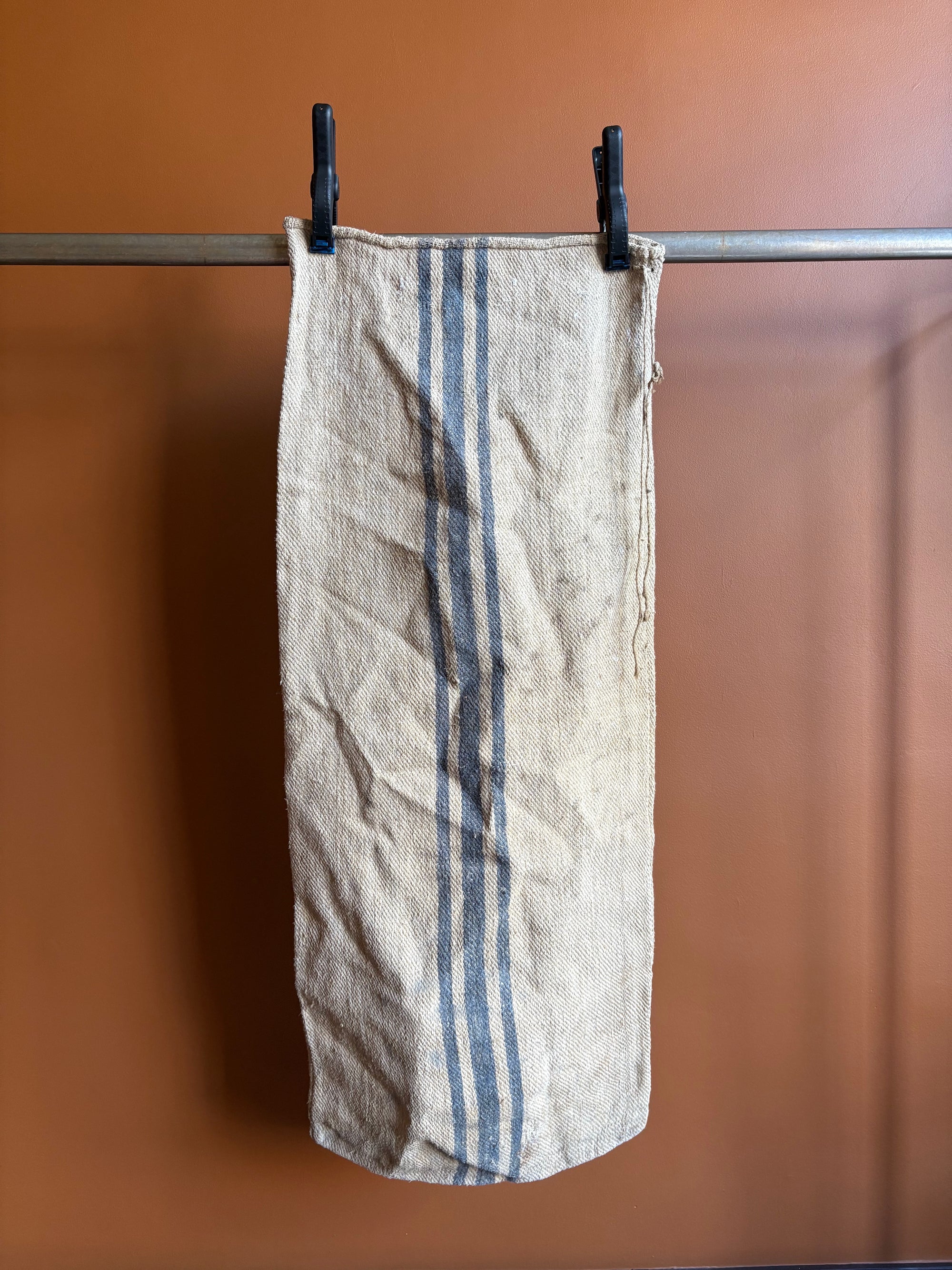 Vintage European burlap grain sack with blue vertical stripes hanging on a metal rail against a terracotta wall.