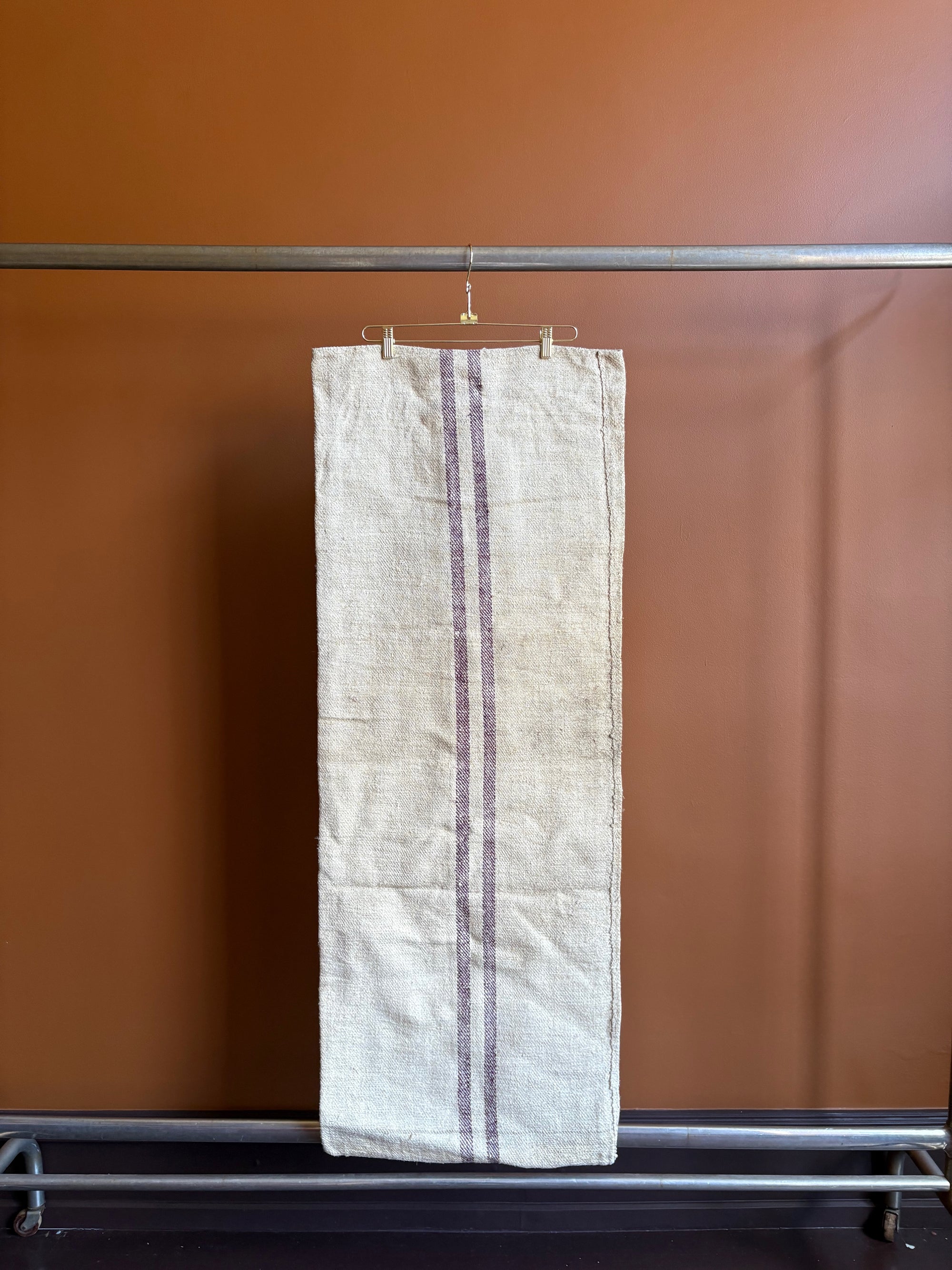 Vintage European burlap grain sack with burgundy purple vertical stripes hanging on a hanger against a terracotta wall.
