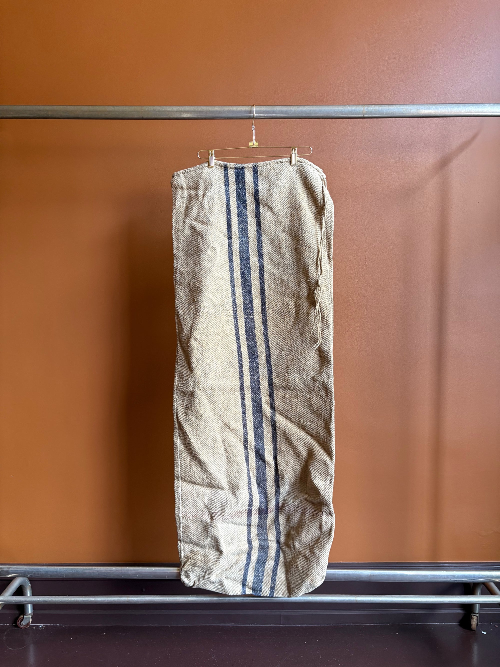 Vintage European burlap grain sack with dark blue vertical stripes hanging on a hanger against a terracotta wall.