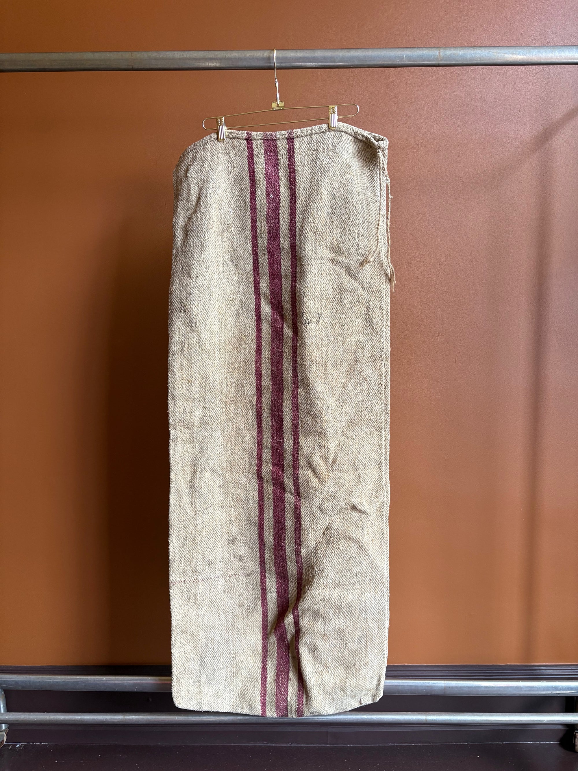 Vintage European burlap grain sack with red burgundy vertical stripes hanging on a hanger against a terracotta wall.