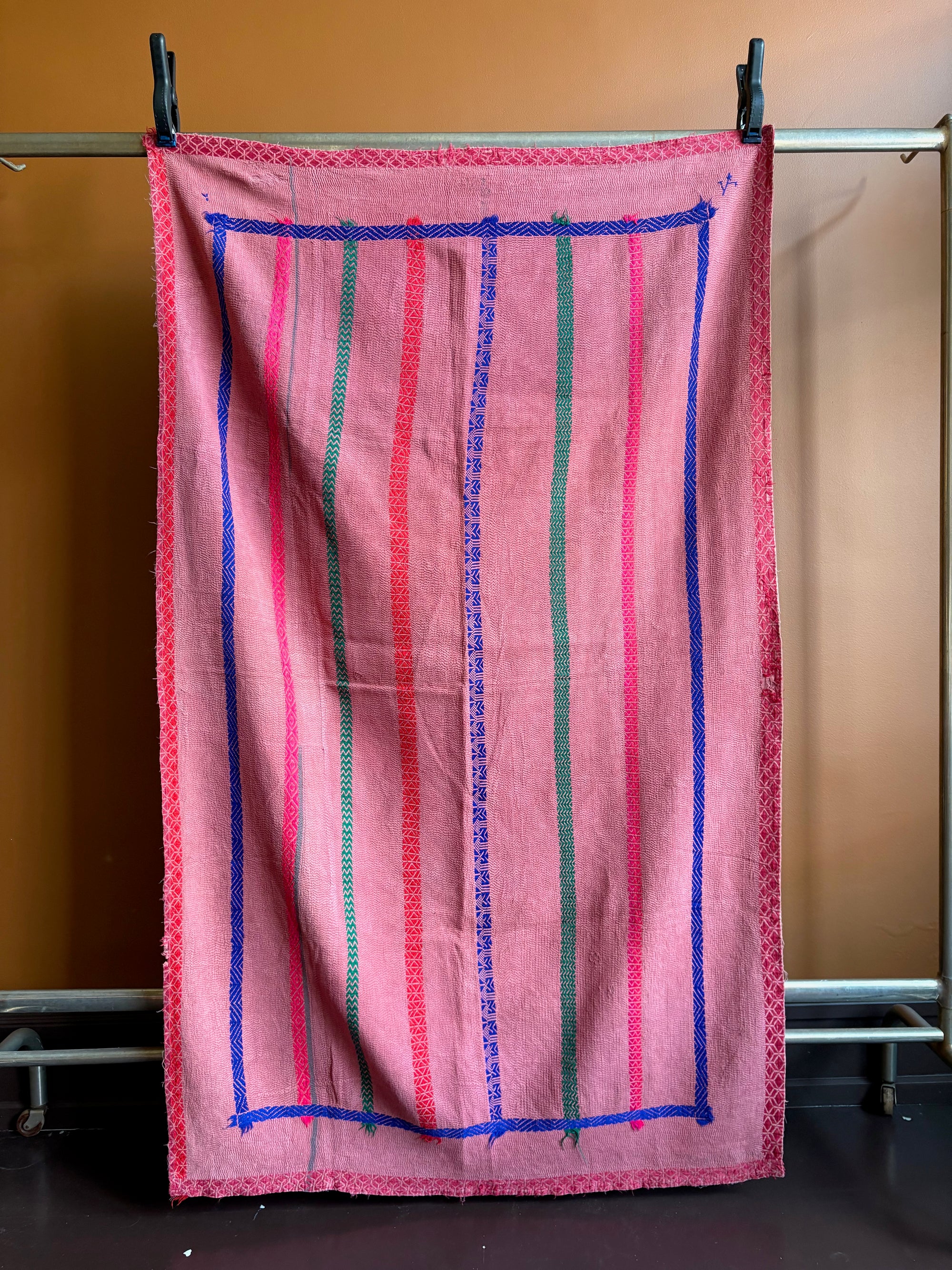 Semi-antique Indian quilt with a warm pink base, contrasting blue border frame, and vertical red and green stripes hanging on a rack against a dark wall.