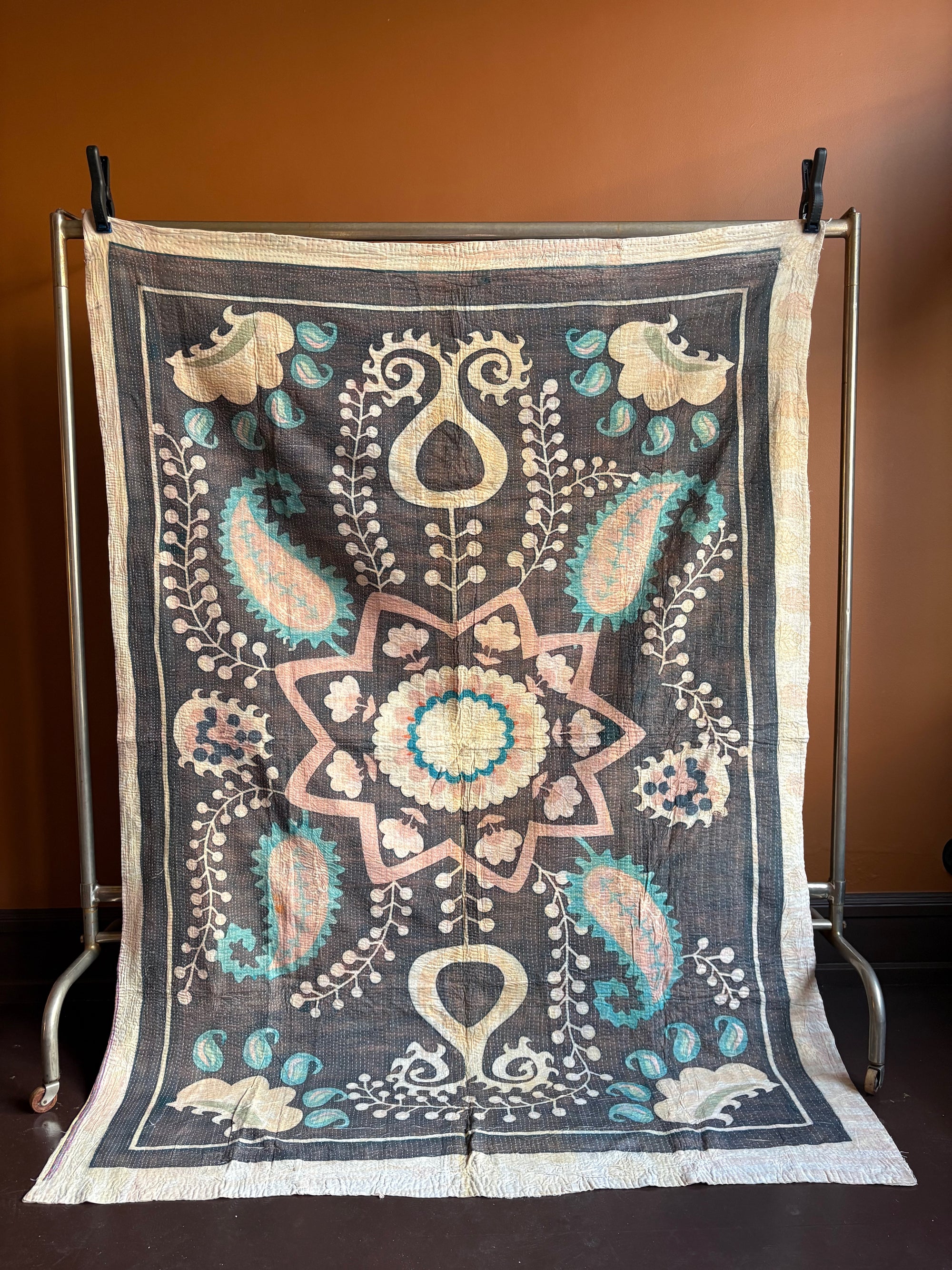 Semi-antique Indian quilt with a charcoal black base, large pink Suzani sun medallion, and turquoise paisley patterns hanging on a rack against a dark wall.