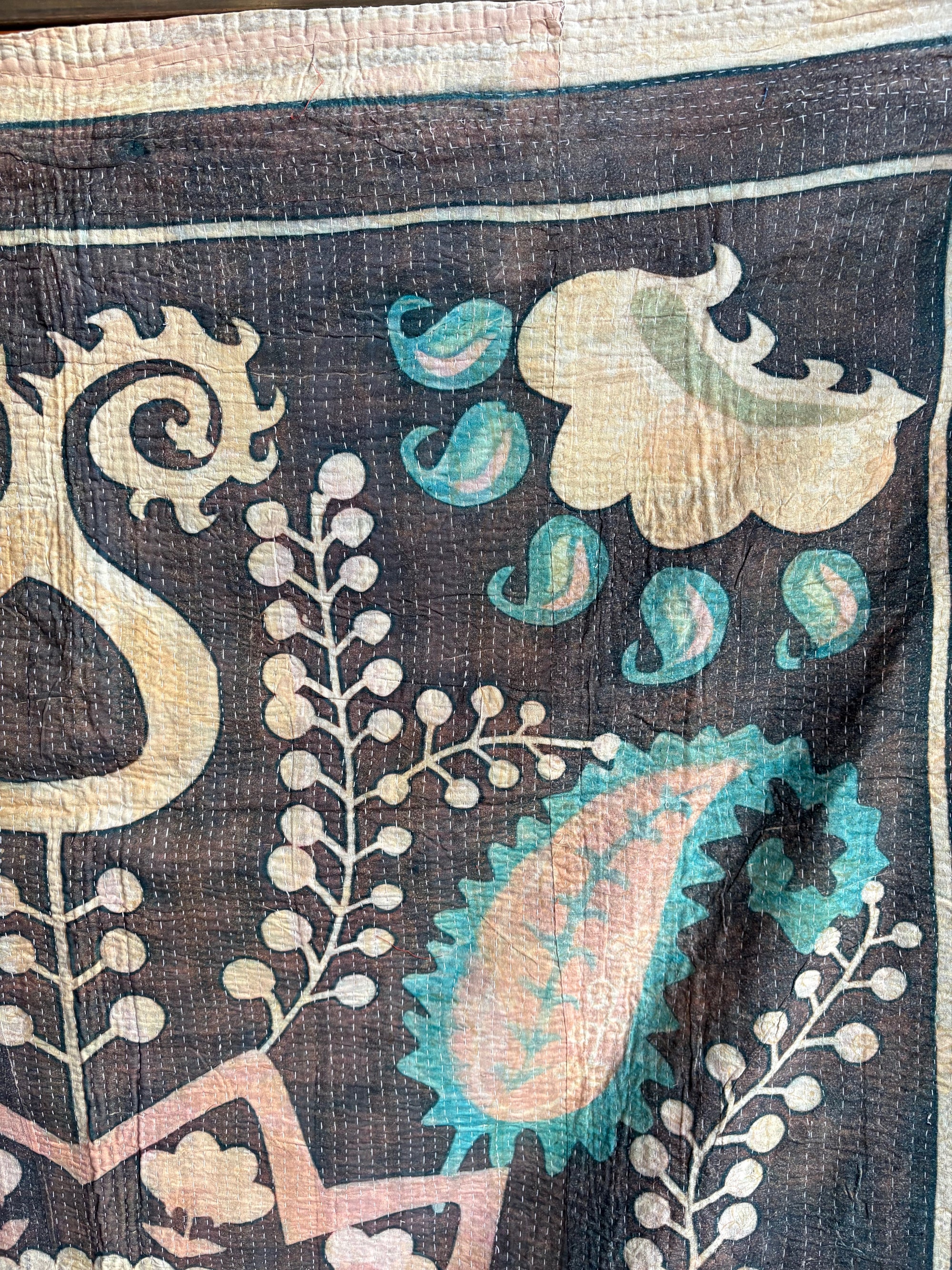 Semi-antique Indian quilt with a charcoal black base, large pink Suzani sun medallion, and turquoise paisley patterns hanging on a rack against a dark wall.