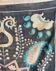 Semi-antique Indian quilt with a charcoal black base, large pink Suzani sun medallion, and turquoise paisley patterns hanging on a rack against a dark wall.