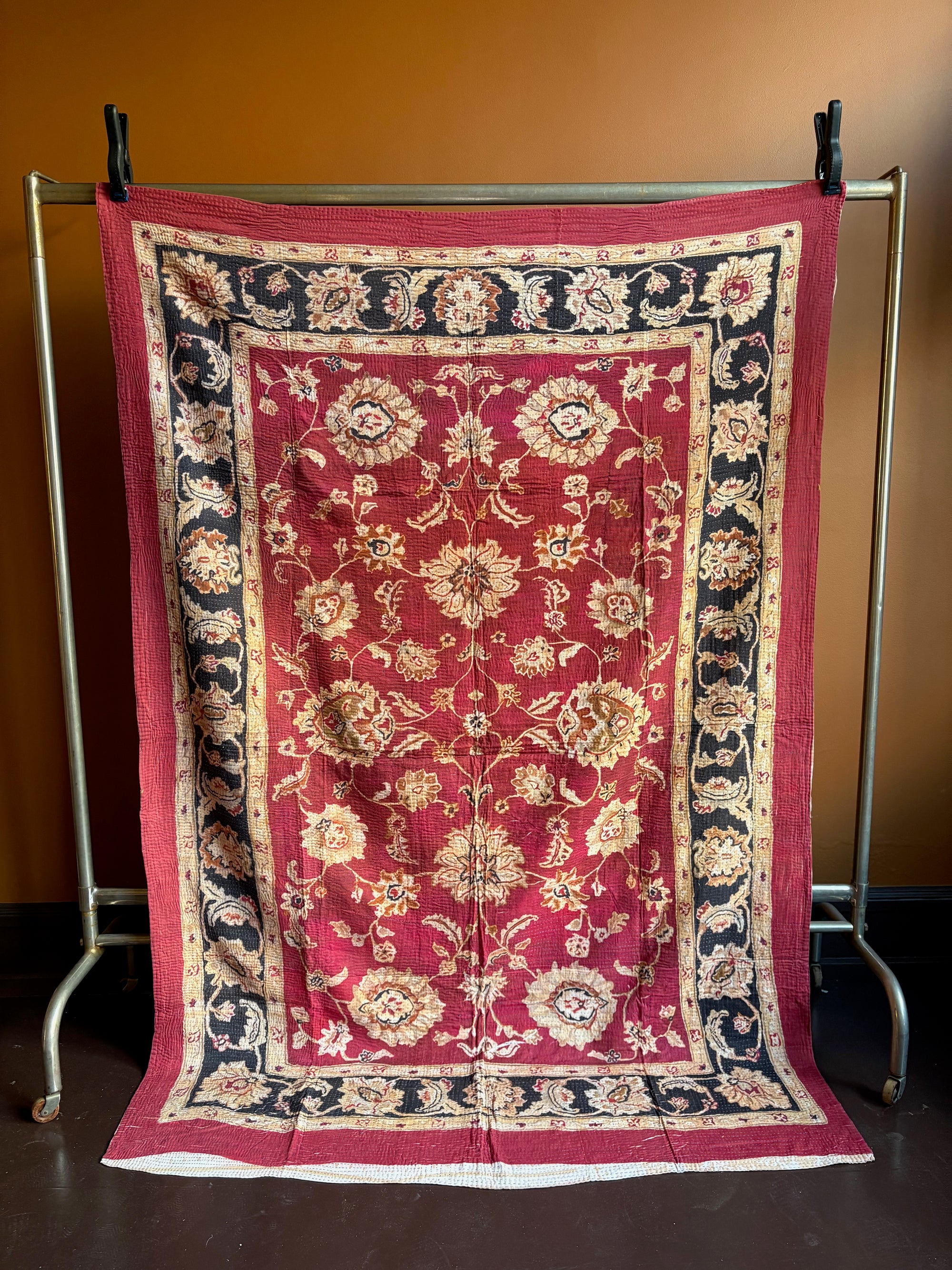 Semi-antique Indian quilt with a deep rust red base, ornate cream floral motifs, and a dark blue patterned border hanging on a rack against a brown wall.