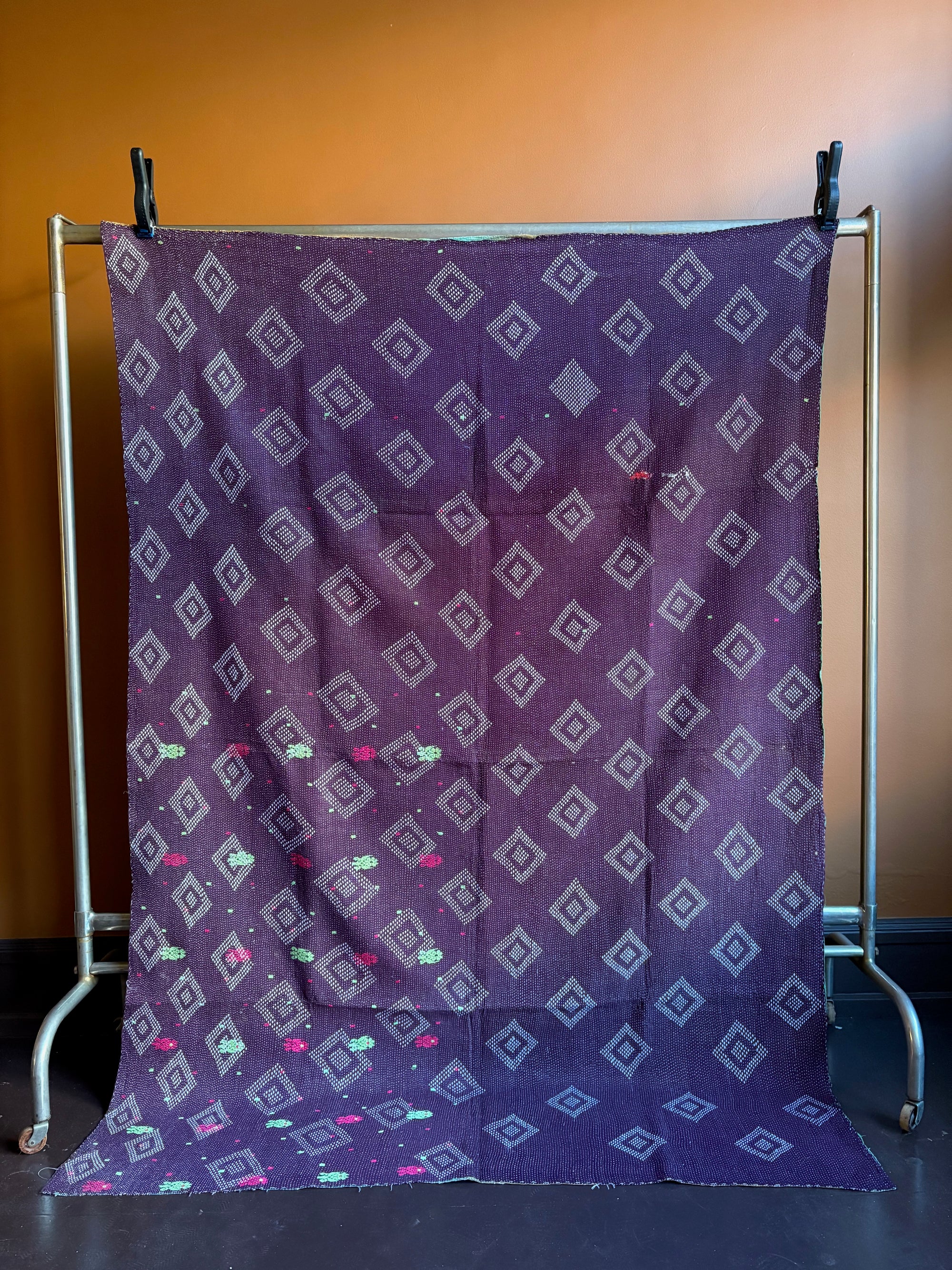 Semi-antique Indian quilt with a deep plum purple base and a white geometric diamond grid pattern hanging on a clothing rack against a brown wall.