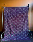 Semi-antique Indian quilt with a deep plum purple base and a white geometric diamond grid pattern hanging on a clothing rack against a brown wall.